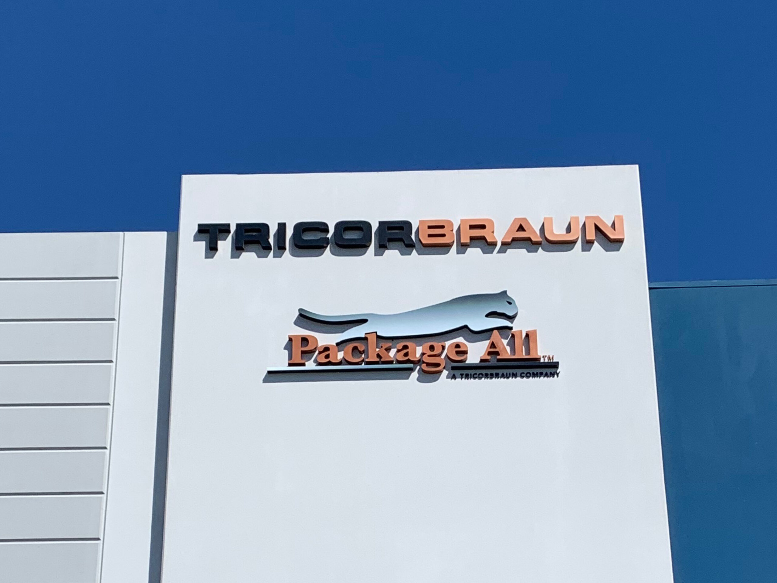 White Projecting Tricorbraun And Package All Lettering Mounted On A Blue Wall Panel.