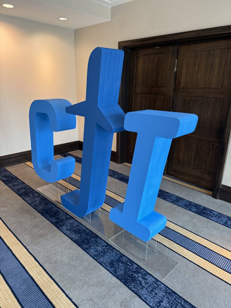 Large Blue Cji Foam Display Letters On Clear Bases Beside Dark Wooden Doors In A Carpeted Event Room.