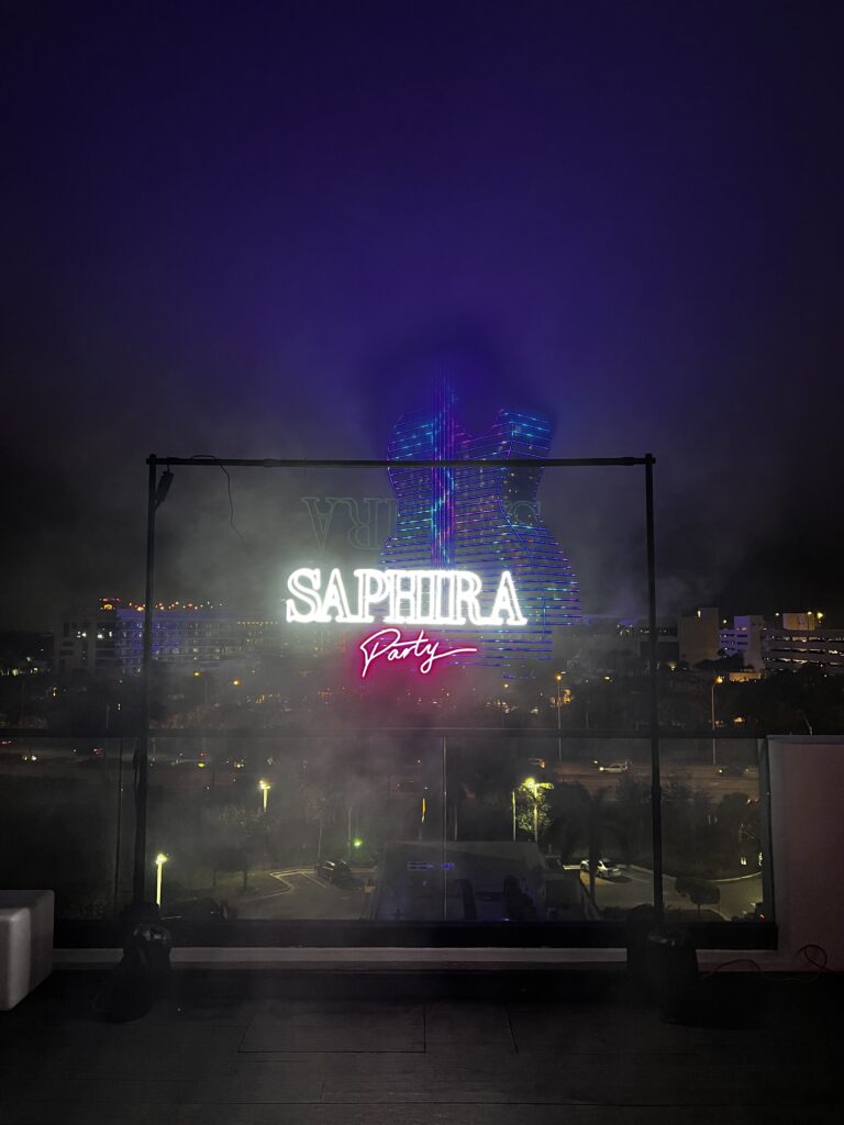 White Saphira Lettering And Pink Party Script Glowing On A Transparent Panel Against A Nighttime City View.