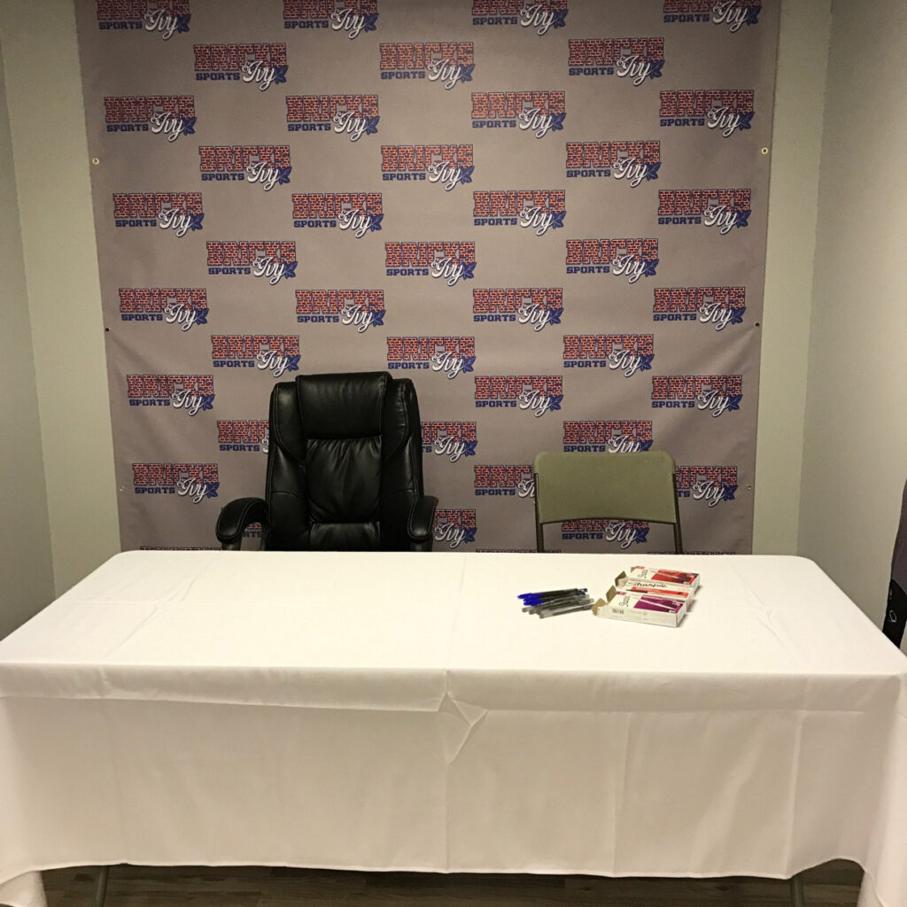 Repeated Red-And-Blue Sports Logos On A Step-And-Repeat Backdrop Behind A White Table And Two Chairs.