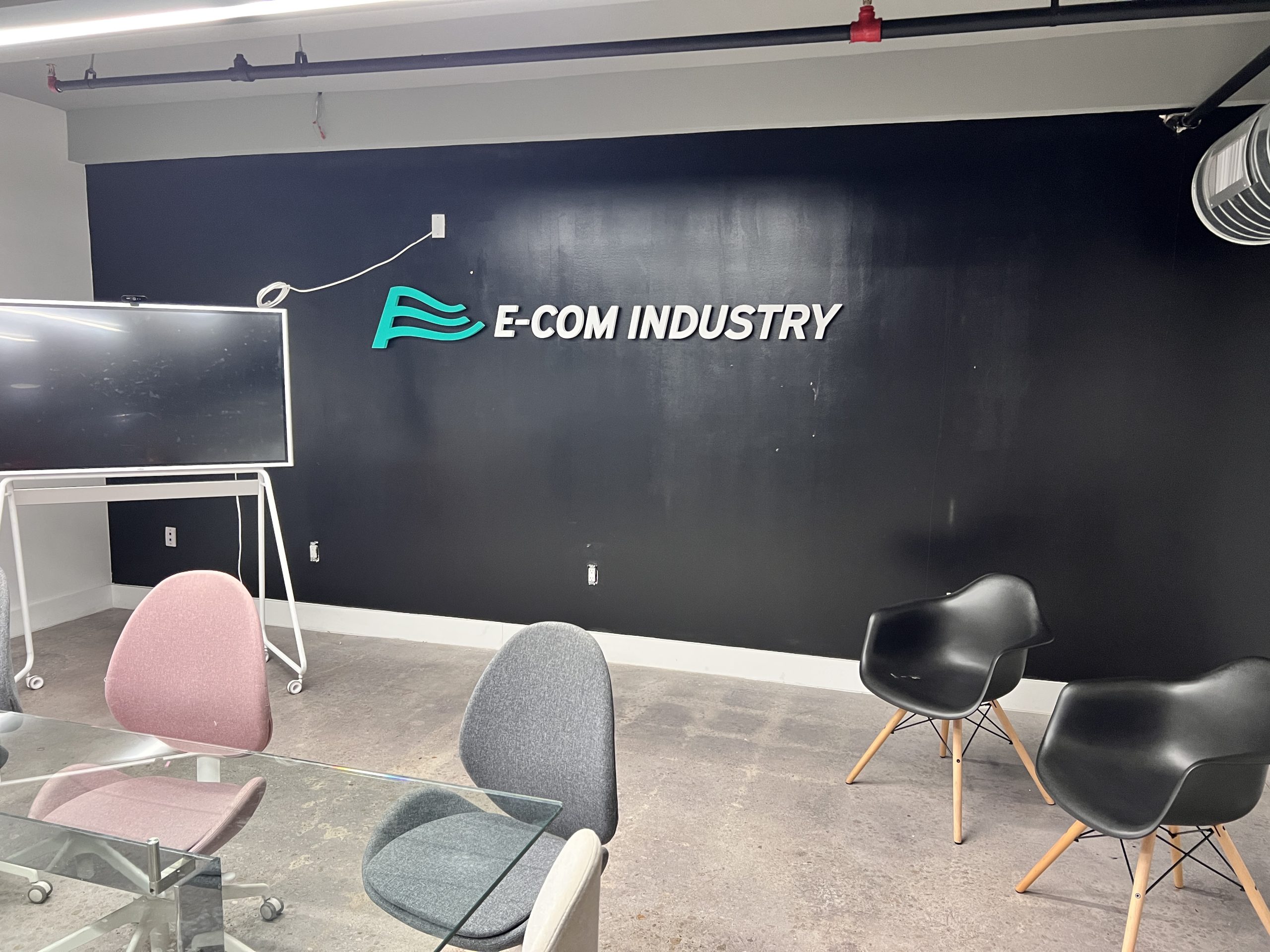 Doral Office Signage And Frosted Glass Graphics | E-Com Industry Raised E-Com Industry Logo And Letters On A Dark Wall In An Office Meeting Area