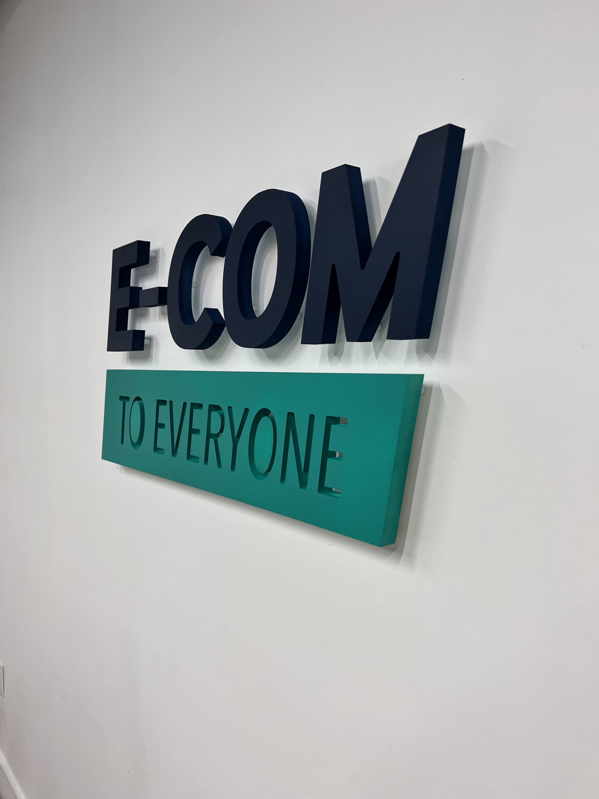 Doral Office Signage And Frosted Glass Graphics | E-Com Industry Raised Dark E-Com Letters Above A Turquoise To Everyone Panel On A White Office Wall