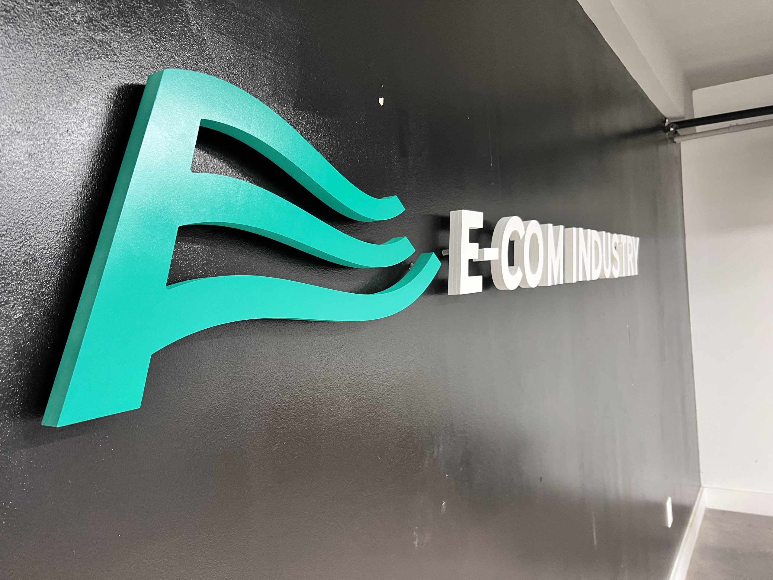 Doral Office Signage And Frosted Glass Graphics | E-Com Industry Turquoise Wave Logo And Raised White E-Com Industry Letters Installed On A Dark Office Wall
