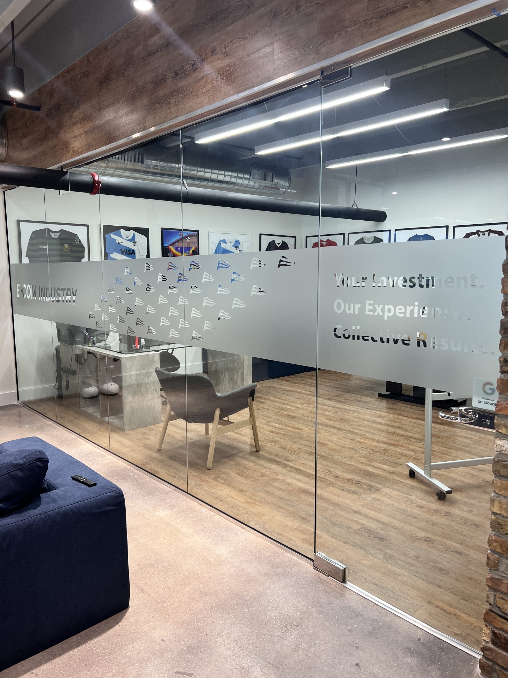 Doral Office Signage And Frosted Glass Graphics | E-Com Industry Long Office Glass Partition With Frosted E-Com Lettering, Repeated Logo Marks, And Framed Shirts Visible Behind It