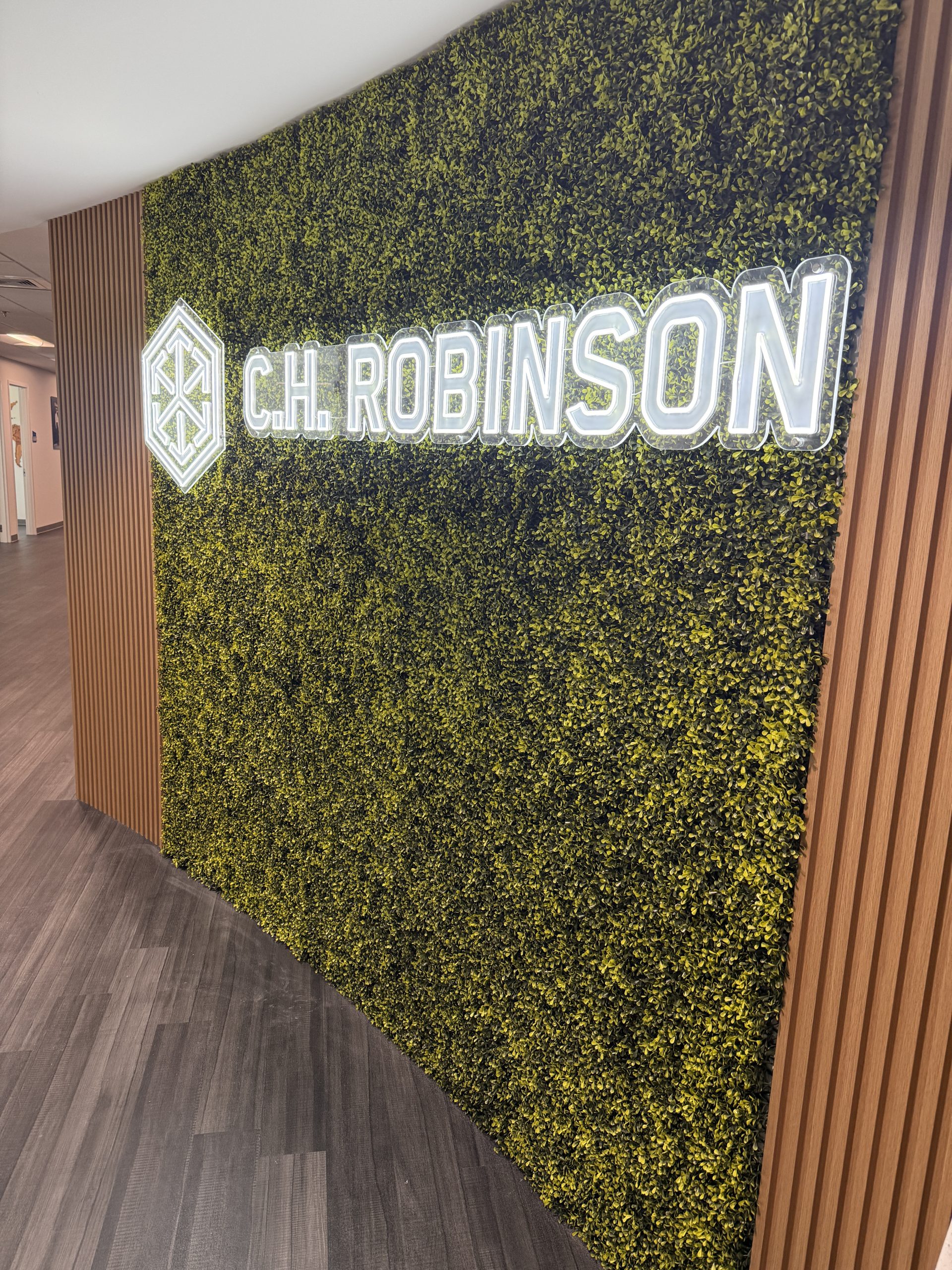 Illuminated LED neon office logo sign mounted on a clear acrylic panel over a green wall feature in a Doral office